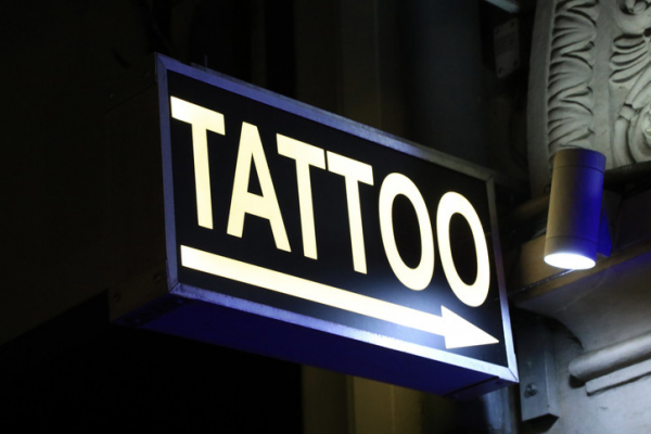 A light shining on a black and dark blue sign that says "Tatooo" in white letters and has an arrow pointing to a doorway